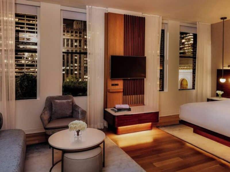Stylish hotel room at The Bryant Park Hotel featuring hardwood floors, modern furnishings, flat-screen TV, and large windows with city views.