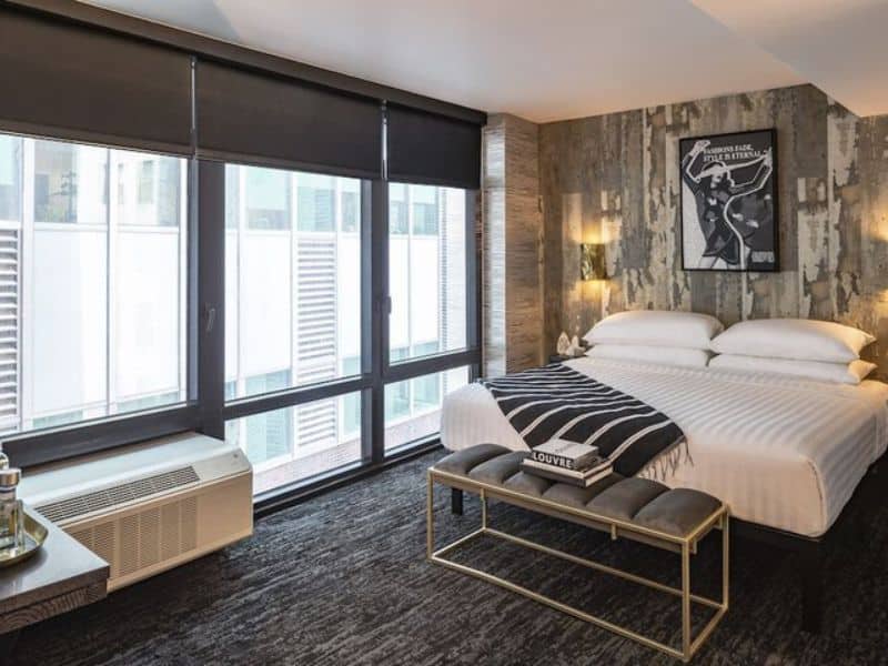 Stylish hotel room at The FIDI Hotel featuring modern decor, chic lighting, plush bedding, and a view through large windows, embodying luxury and comfort in Manhattan's Financial District.