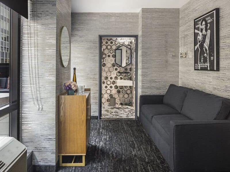 Stylish hotel room at The FIDI Hotel featuring modern decor, chic lighting, plush sofa, and a view of the bathroom with hexagonal tile design.