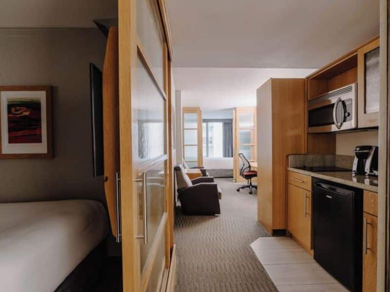 Modern hotel suite interior at The Jewel Hotel, New York, featuring kitchenette with microwave, refrigerator, and coffee maker, workspace, and seating area with natural light from windows.
