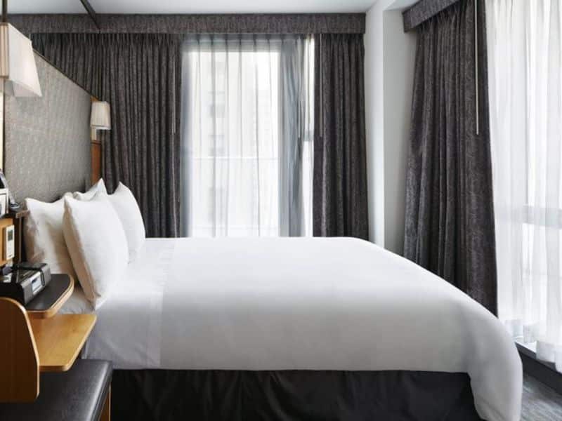 Modern hotel room at The Jewel Hotel, New York, featuring a queen bed, stylish decor, floor-to-ceiling windows with sheer curtains, and bedside table with amenities.
