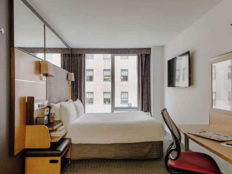 Modern hotel room at The Jewel Hotel, New York, featuring a queen bed, flat-screen TV, workspace, and large windows with city views.