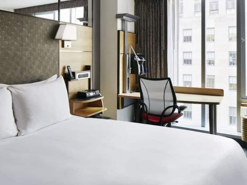 Modern hotel room at The Jewel Hotel, New York, featuring a comfortable bed, workspace with desk and chair, and large windows allowing natural light.