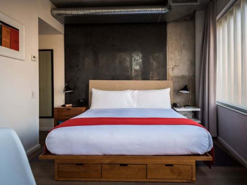 Stylish hotel room at The Nolitan featuring a modern bed with a red accent, hardwood floors, and large windows, emphasizing comfort and contemporary design.