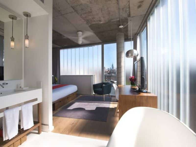 Stylish hotel room at The Nolitan featuring hardwood floors, large windows with city views, modern furnishings, and a luxurious freestanding bathtub.