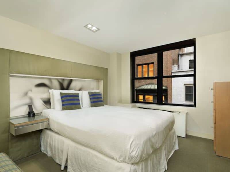Stylish hotel room at The Shoreham Hotel in Midtown Manhattan, featuring a queen bed with blue striped pillows, minimalist decor, and large window with urban view.