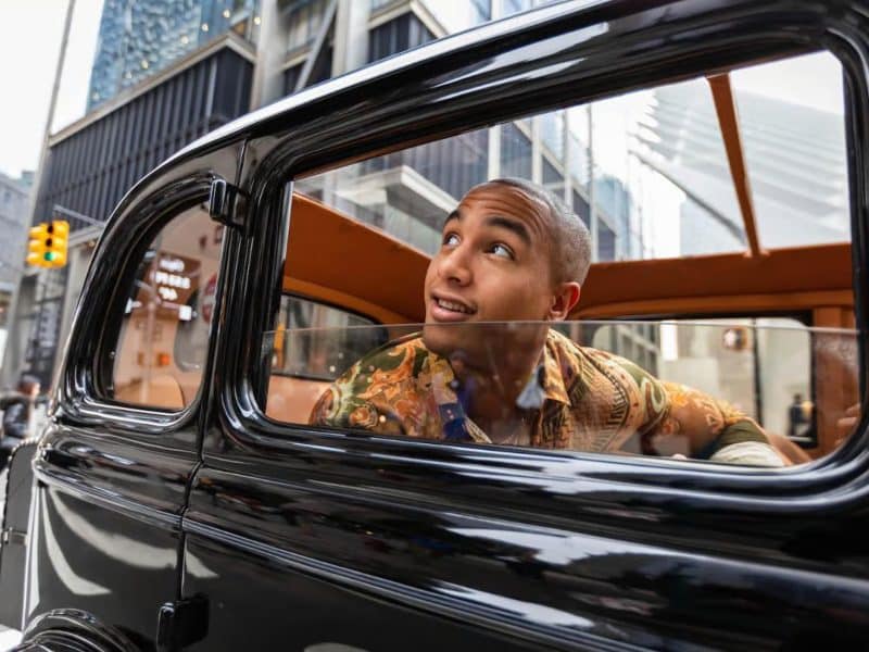 Person enjoying a scenic city tour in a vintage car, smiling and looking out the window, with urban architecture in the background, representing the Vintage Wine Tasting and Classic Car Combo Tour experience.