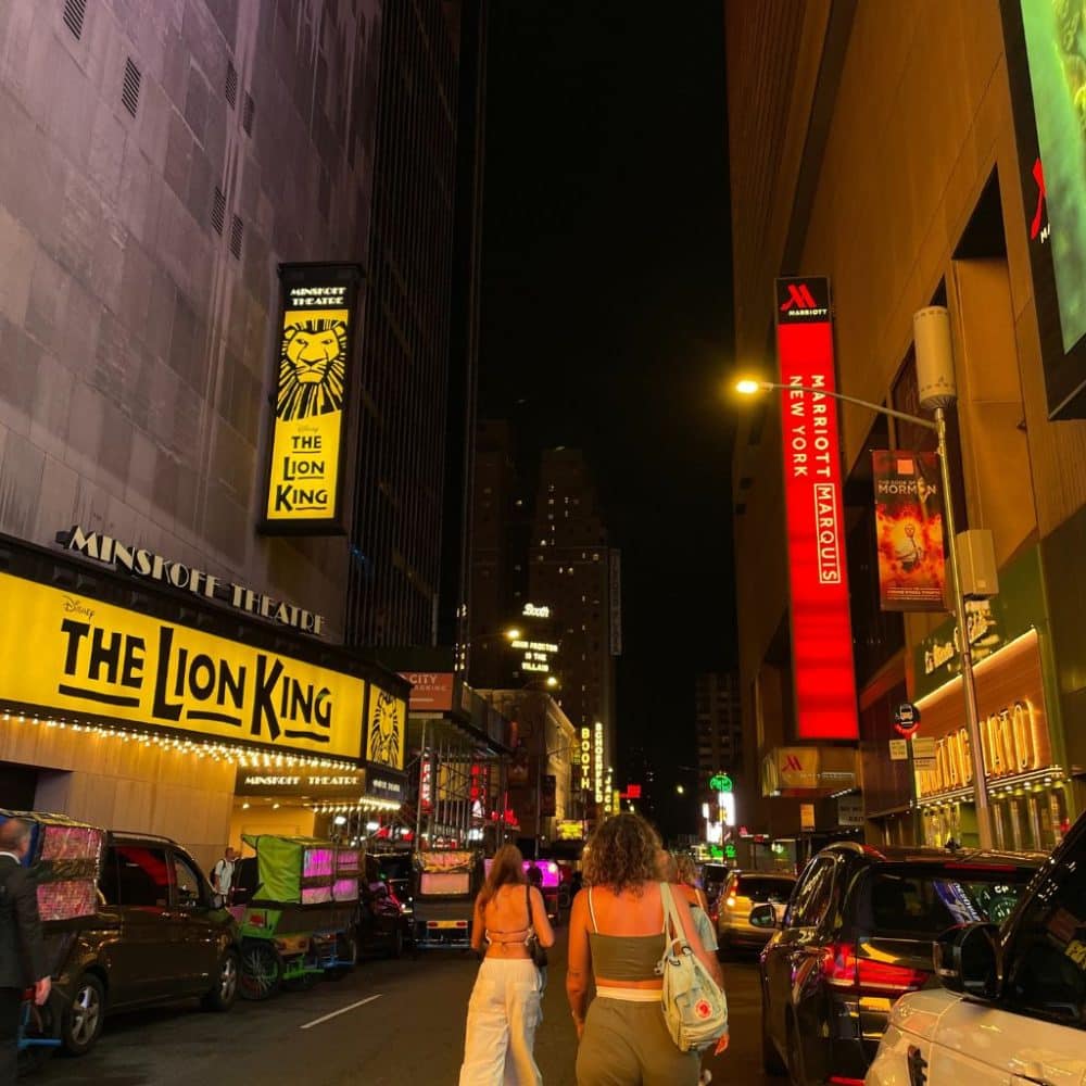 Times Square at night featuring the bright marquee of "The Lion King" at the Minskoff Theatre, pedestrians walking, and illuminated buildings including the Marriott Marquis.