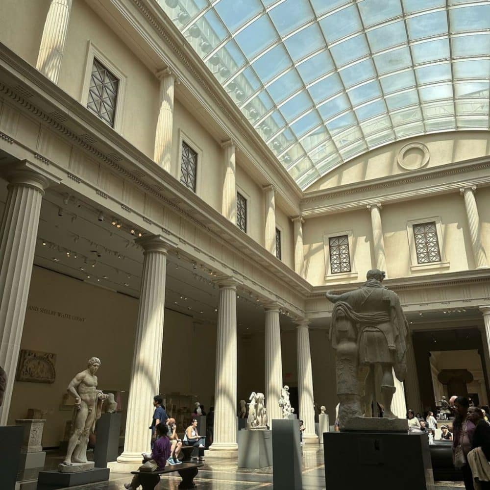 Interior view of The Met museum showcasing classical sculptures, visitors exploring exhibits, and natural light streaming through a glass ceiling.