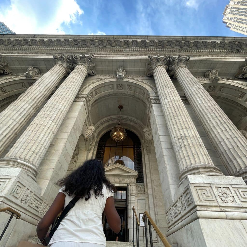 Person ascending the steps of the New York Public Library, showcasing its grand architecture with tall columns and intricate details, reflecting a detour experience in NYC.