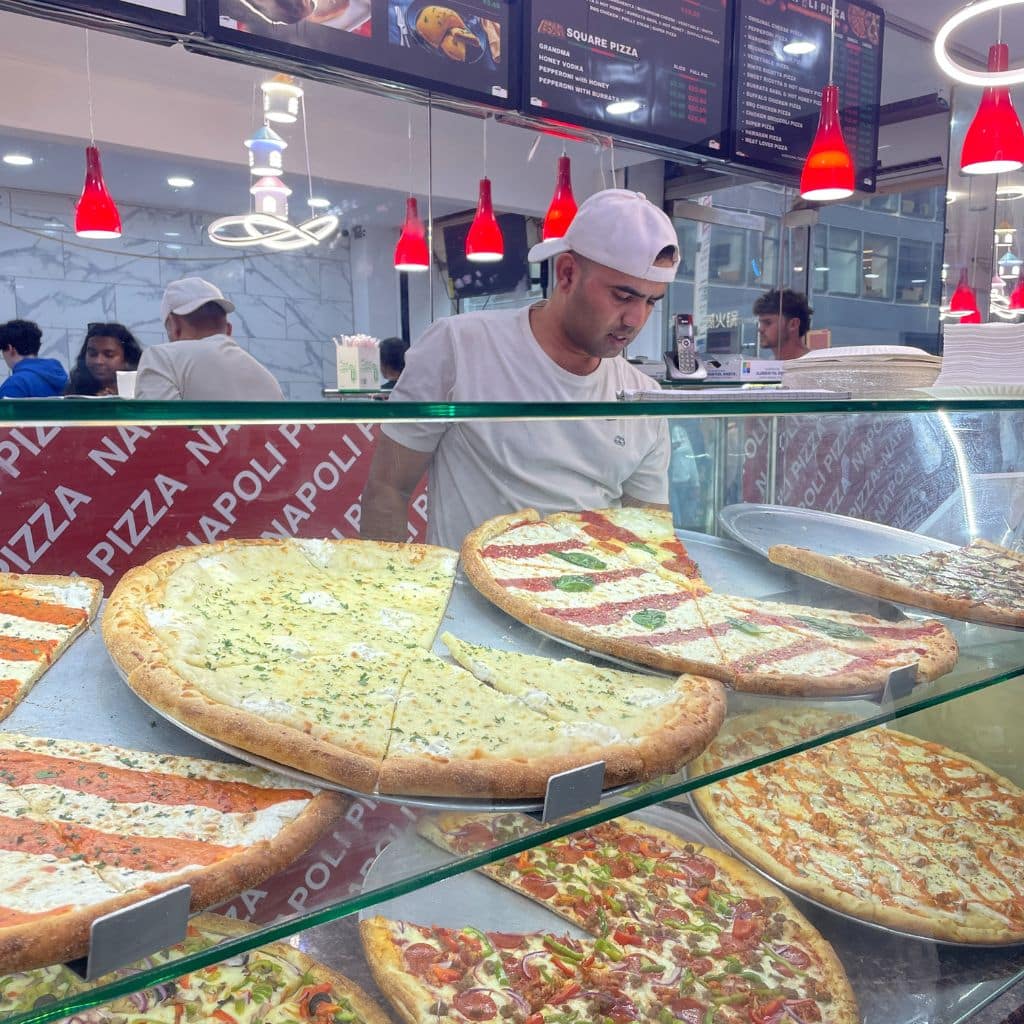 Pizza slices displayed in a glass case at a New York pizzeria, with a staff member in a white shirt and cap serving customers, showcasing various toppings and styles.