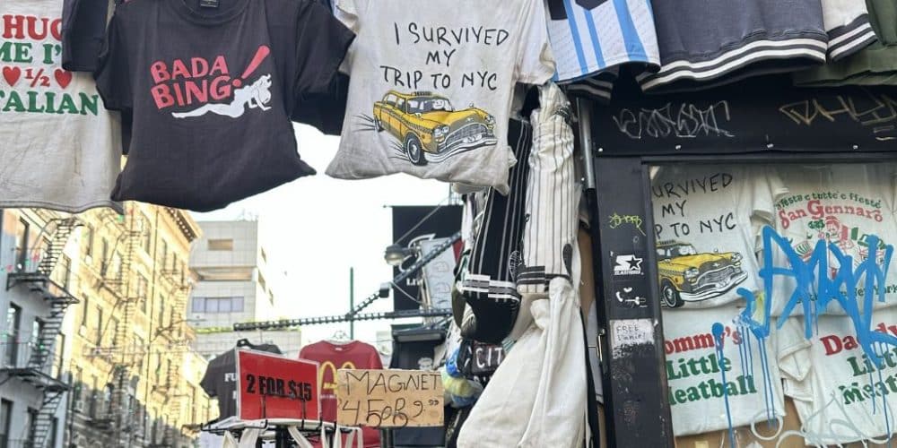 Souvenir NYC t-shirts displayed on a street vendor's rack, featuring phrases like "I Survived My Trip to NYC" and "BADA BING!", capturing the vibrant shopping scene in Little Italy.