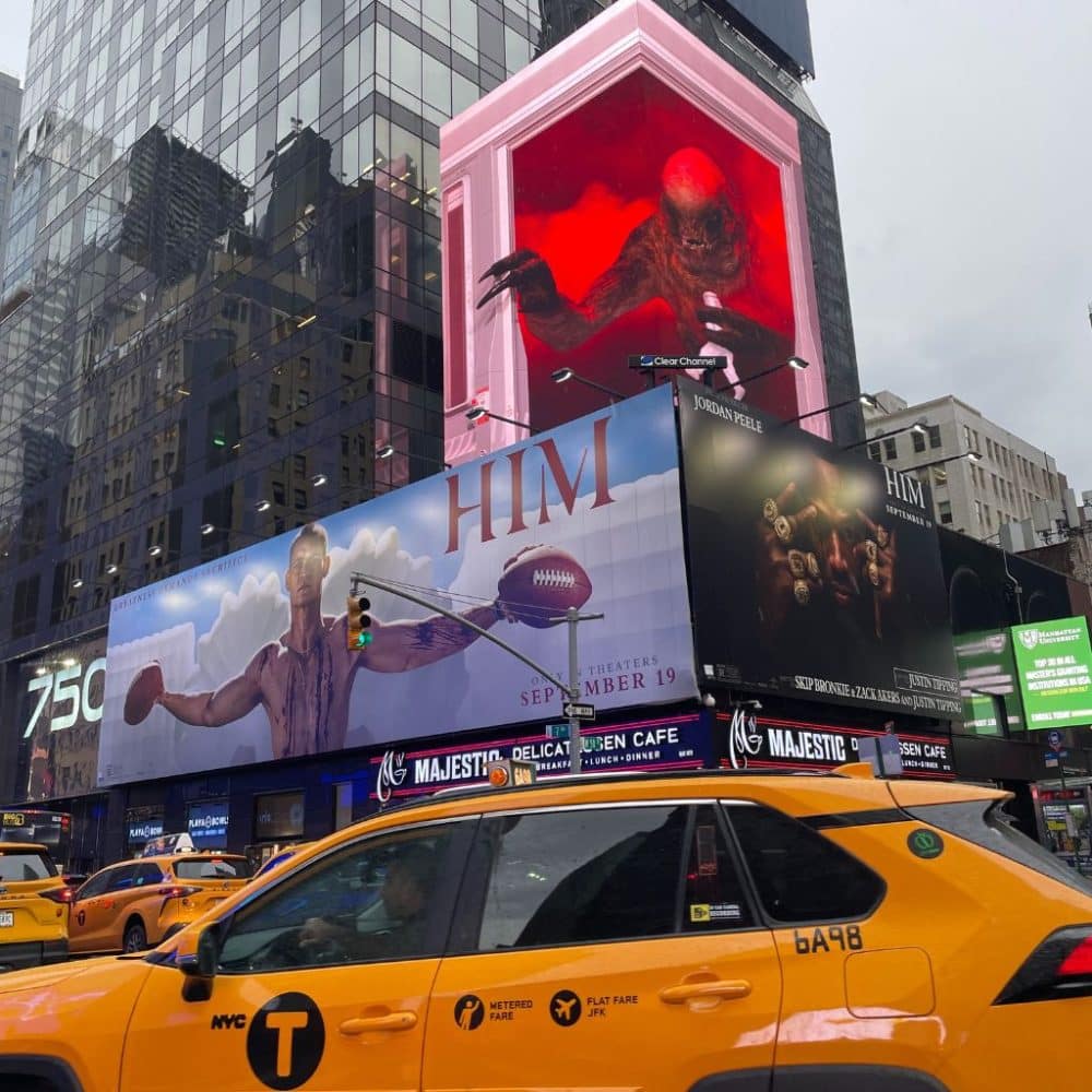 Billboards in Times Square featuring promotional ads for a film titled "HIM," displaying a muscular man with a football and a dramatic creature in a red-lit box, with yellow NYC taxis in the foreground.
