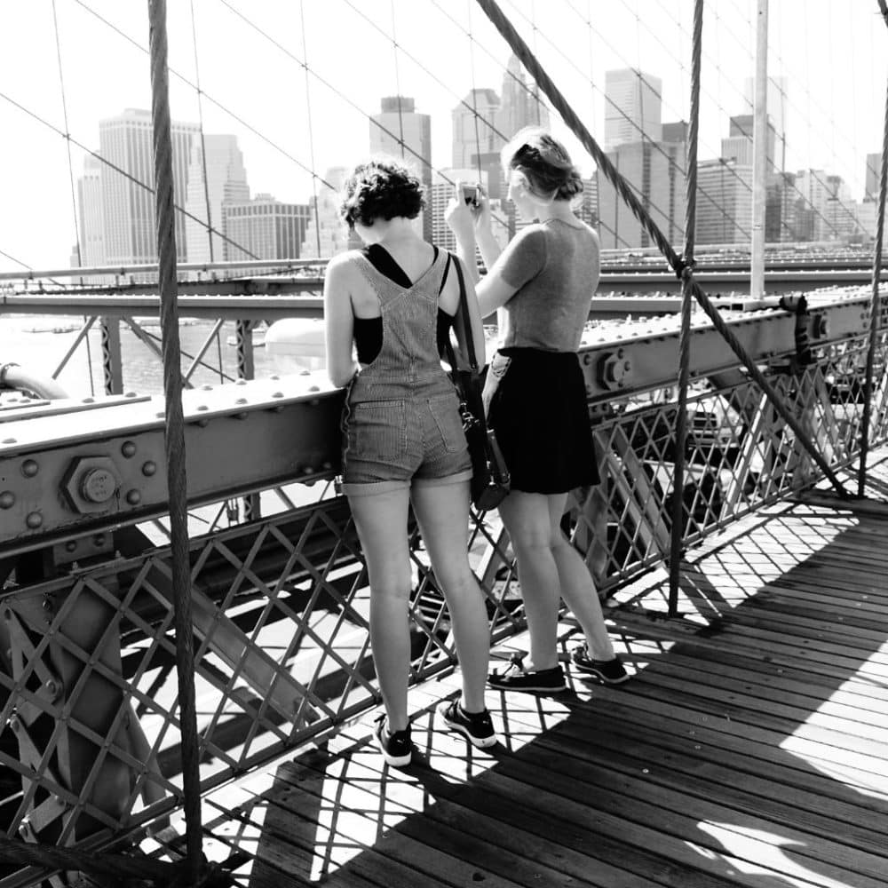 Two women in comfortable summer outfits, one in overalls and the other in a skirt, enjoying the view of the New York City skyline from a bridge, emphasizing practical summer clothing for city exploration.