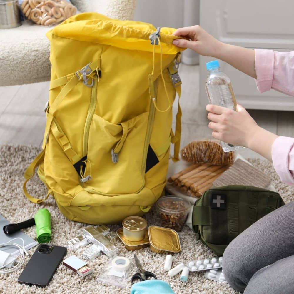 Yellow backpack with open top, person holding water bottle, surrounded by travel essentials like snacks, toiletries, and tech gear, illustrating practical packing for a summer trip to New York City.