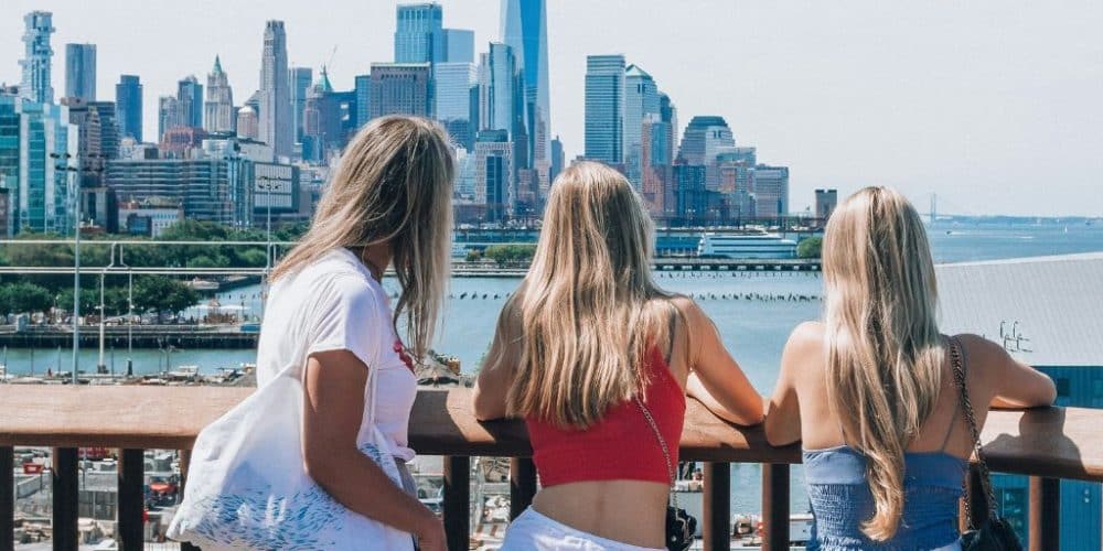 Three women enjoying a summer view of the New York City skyline, overlooking the waterfront with modern buildings and a clear sky in the background.