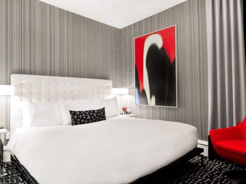 Modern hotel room at Moderne Hotel, featuring a plush white bed with black accent pillows, contemporary artwork on the wall, and a stylish red chair, highlighting the hotel's boutique and pet-friendly atmosphere.