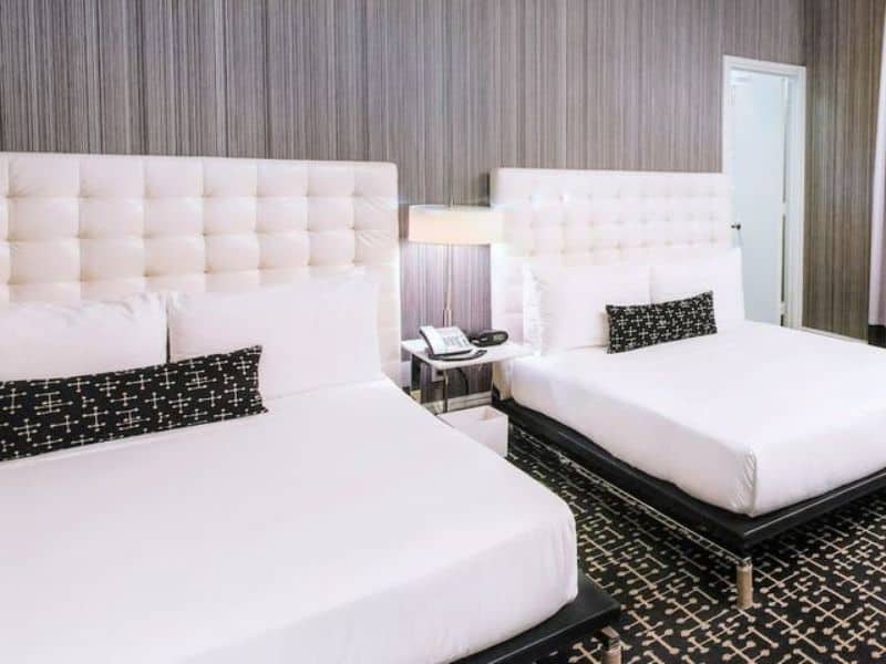 Modern hotel room with two beds, stylish white headboards, contemporary decor, bedside lamp, phone, and dark patterned carpet, representing the Moderne Hotel's accommodation features in New York City.