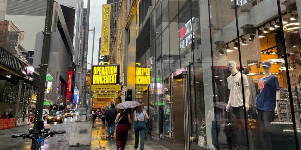 Rainy NYC street scene in Hell's Kitchen with pedestrians under umbrellas, vibrant theater signage for "Operation Mincemeat," and reflections on wet pavement, capturing the urban atmosphere and diverse experiences of the city.