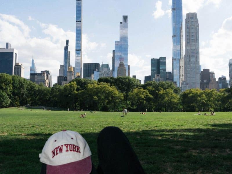 Person relaxing on grassy field in Central Park, wearing a "New York" cap, with NYC skyline in the background, reflecting on travel experiences and personal growth.