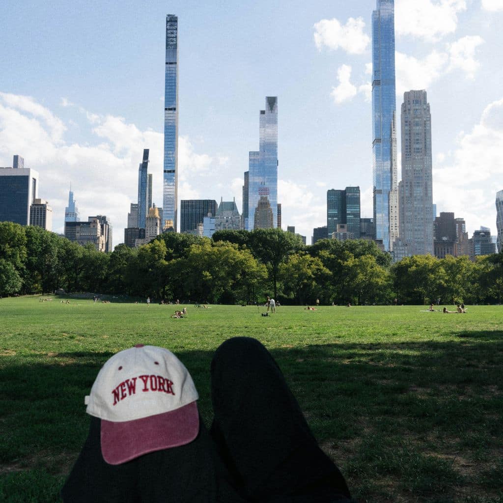 Cap wearing "New York" in foreground with Central Park grass and NYC skyline in background, showcasing iconic cityscape and leisure atmosphere.