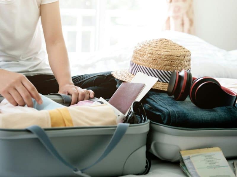 Person packing a suitcase with summer essentials like clothing, a straw hat, headphones, and travel accessories, highlighting practical packing for a New York City trip.