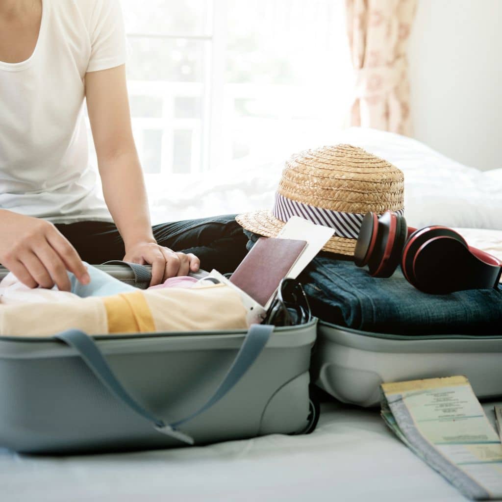 Person packing a suitcase with summer clothing, a straw hat, headphones, and travel documents, preparing for a trip to New York City.