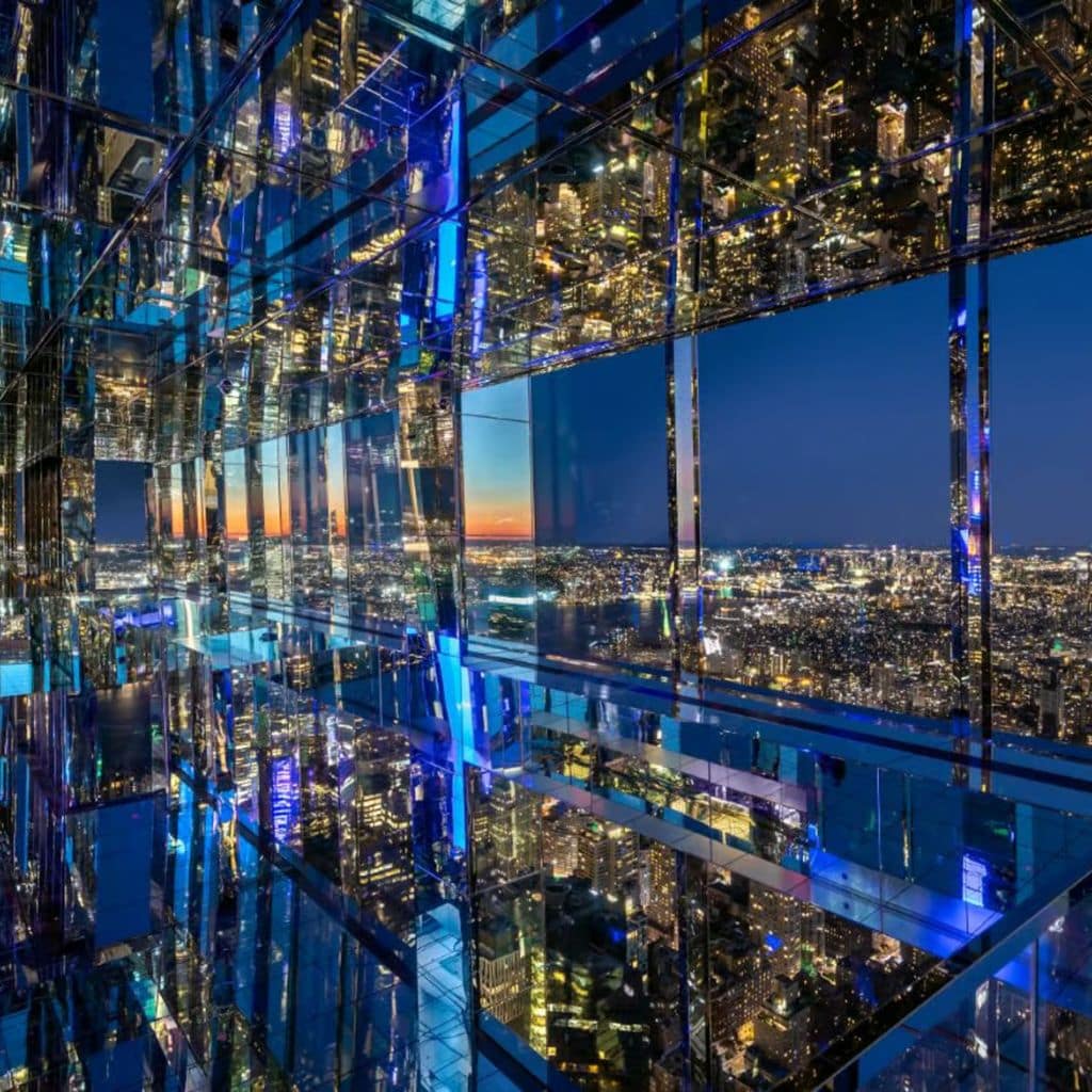 Reflective interior of Summit One Vanderbilt showcasing panoramic views of New York City skyline at sunset, with vibrant city lights and mirrored surfaces enhancing the experience.