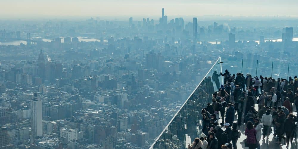 Crowd of visitors at an NYC observation deck overlooking a panoramic view of the city skyline and harbor, highlighting iconic buildings and distant landmarks.