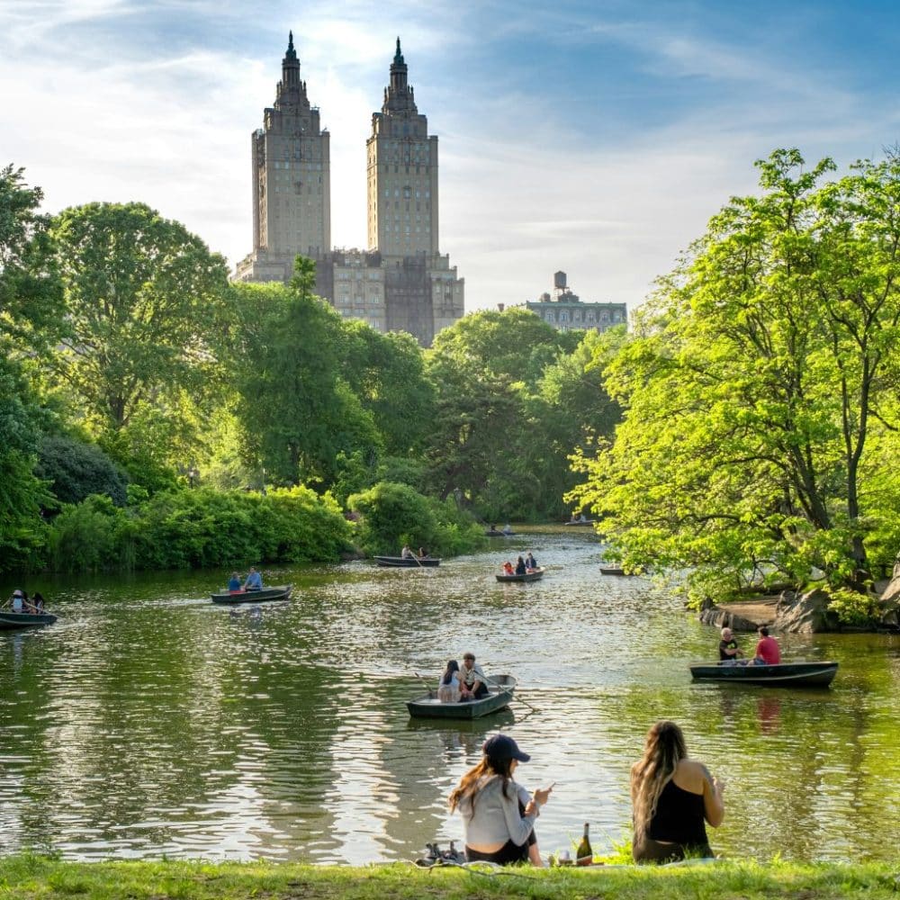 Central Park scene with two women sitting by the water, enjoying a picnic, surrounded by lush greenery and boats on the lake, featuring the iconic twin towers of the San Remo building in the background, reflecting the spirit of spontaneous exploration in NYC.