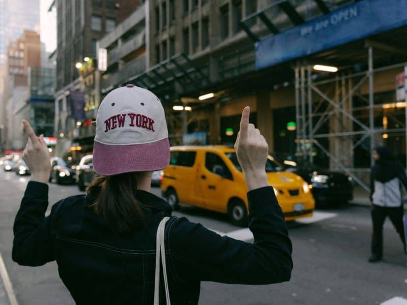 Person wearing a "New York" cap gesturing in a bustling NYC street with yellow cabs and urban buildings, reflecting the excitement of visiting New York City.