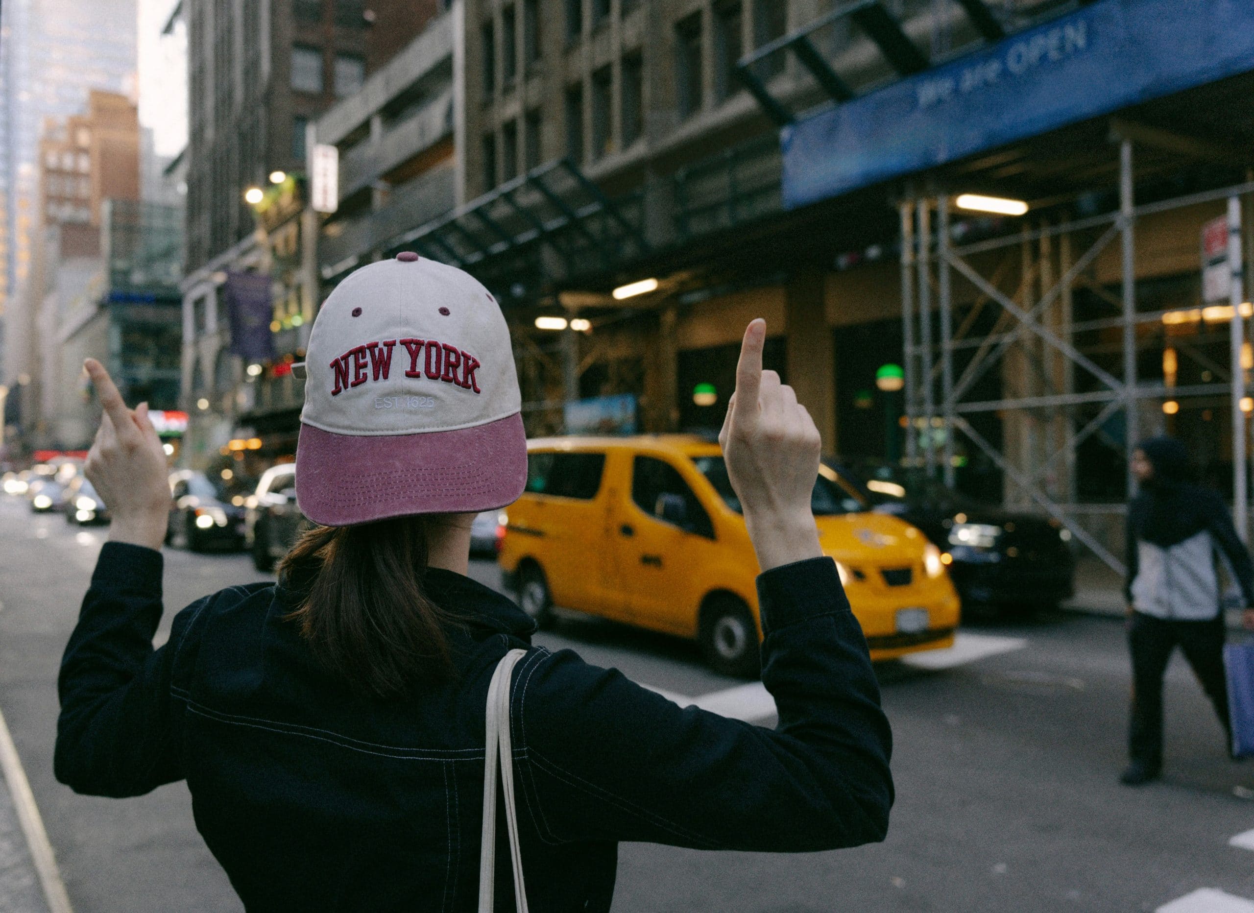 Person wearing a "New York" cap, gesturing in a busy NYC street with yellow taxis and urban buildings in the background, reflecting a visitor's excitement in exploring New York City.