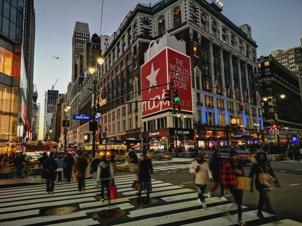 Macy's department store, known as the world's largest store, in a busy New York City street scene with pedestrians crossing at dusk, showcasing vibrant city lights and urban activity.