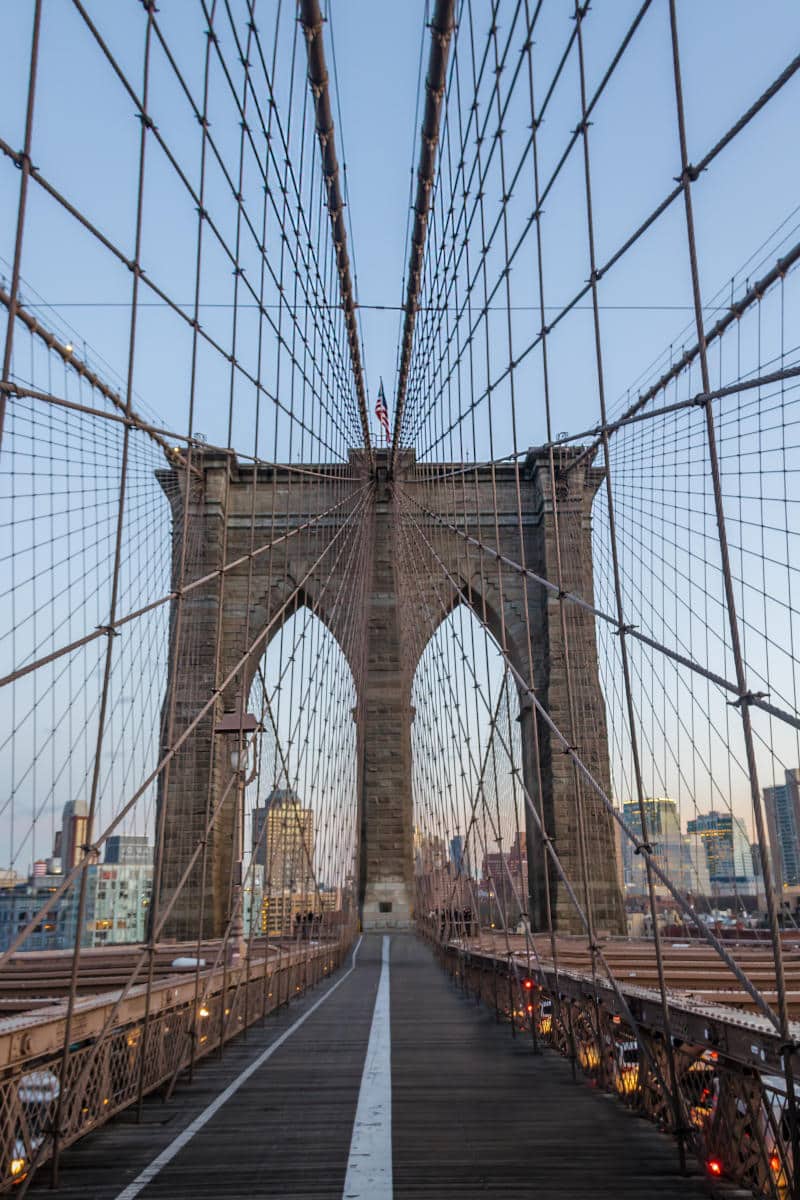 Brooklyn Bridge viewed from the walkway, showcasing the iconic arches and suspension cables against a clear sky, symbolizing New York City's landmarks and attractions.