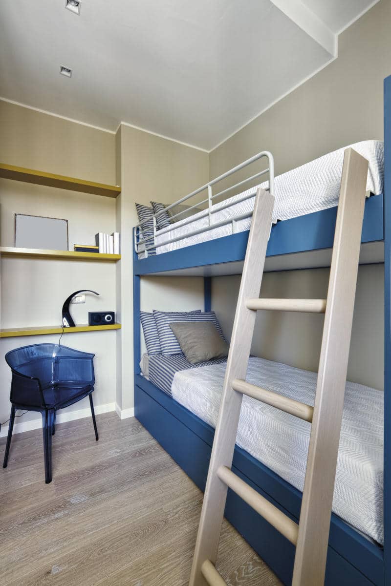 Bunk beds in a modern hotel room with a blue frame, striped bedding, and a wooden ladder, featuring a chair and shelves, suitable for family-friendly accommodations in New York City.