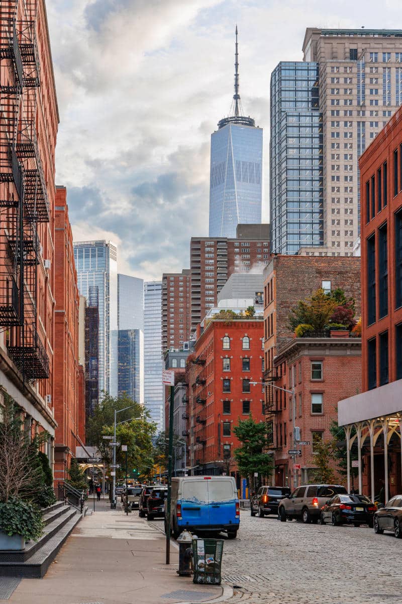 Lower Manhattan street scene featuring red brick buildings, modern skyscrapers including One World Trade Center, and parked vehicles, illustrating NYC's architectural blend.