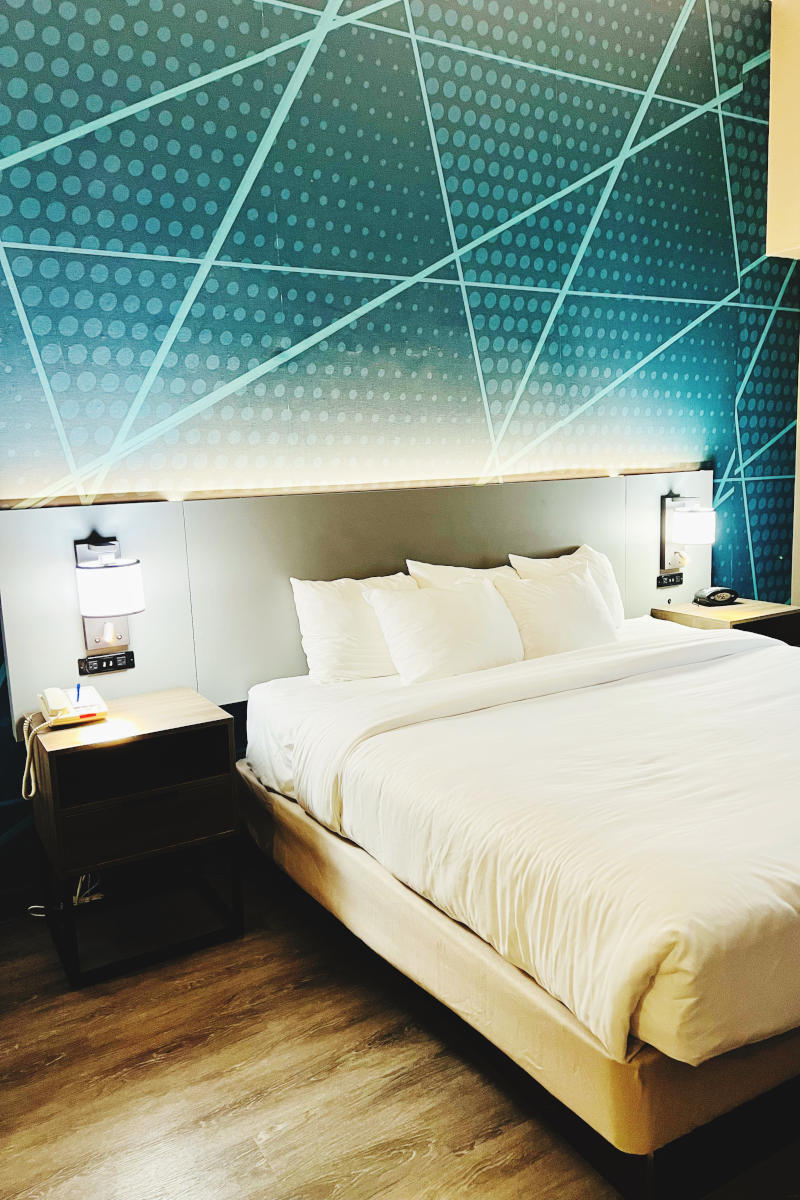 Modern hotel room featuring a queen bed with white linens, stylish blue patterned wall, nightstand with lamp and phone, designed for comfort and convenience in New York City.