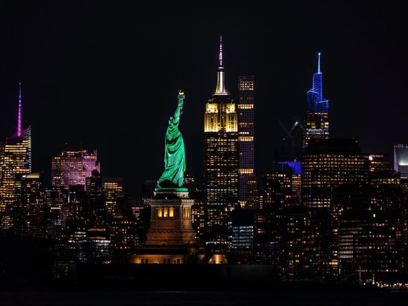 Statue of Liberty illuminated at night with NYC skyline featuring Empire State Building and modern skyscrapers in the background, representing must-see attractions in New York City.