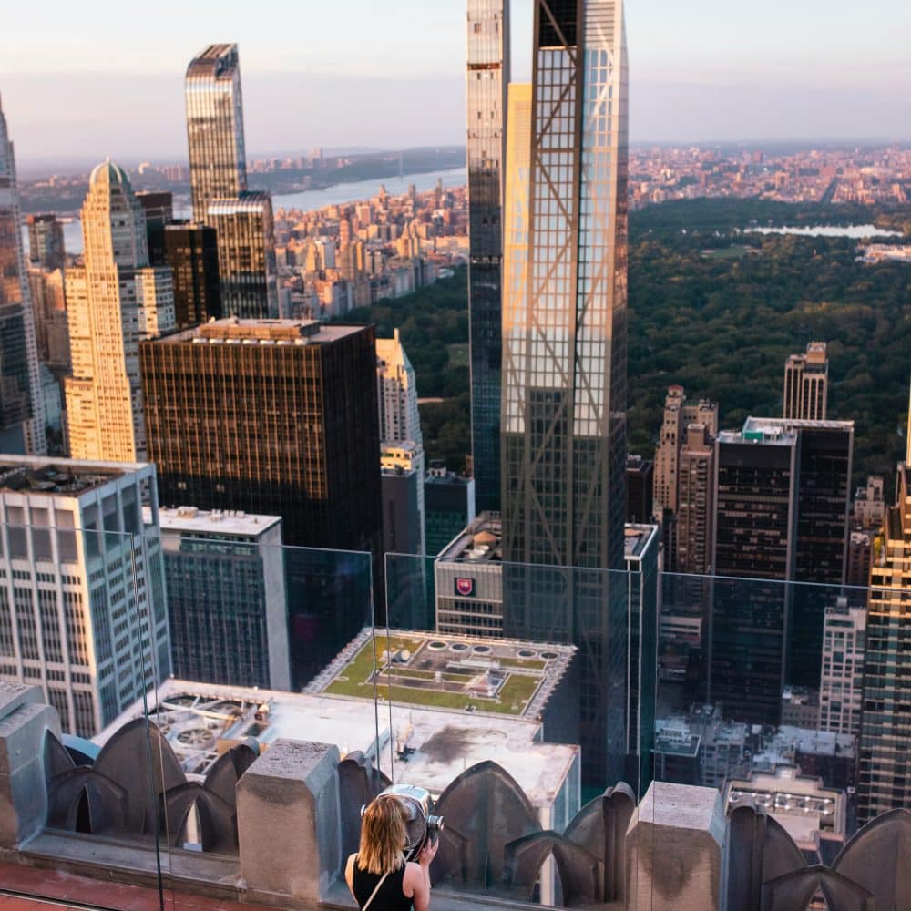 View from Top of the Rock observation deck showcasing Central Park, Midtown Manhattan skyline, and a visitor taking photographs during golden hour.