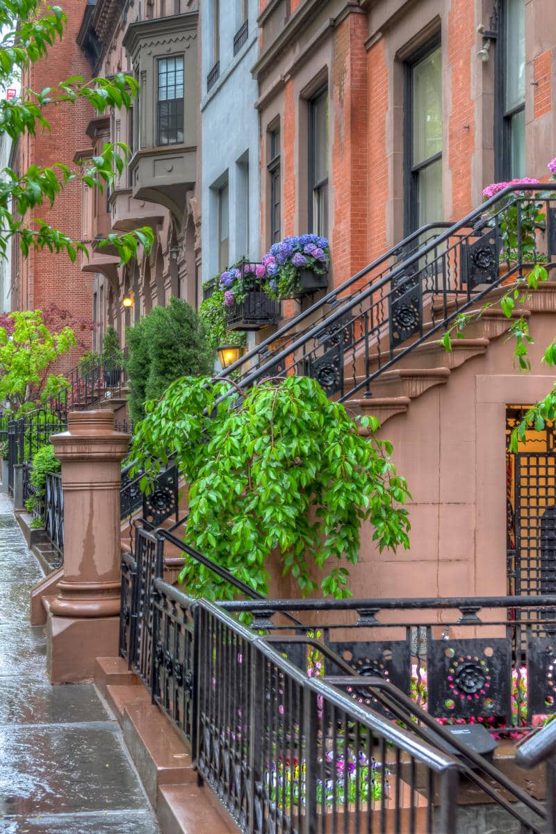 Residential brownstone buildings in Upper Manhattan, featuring lush greenery, colorful flower boxes, and ornate iron railings along a wet sidewalk.